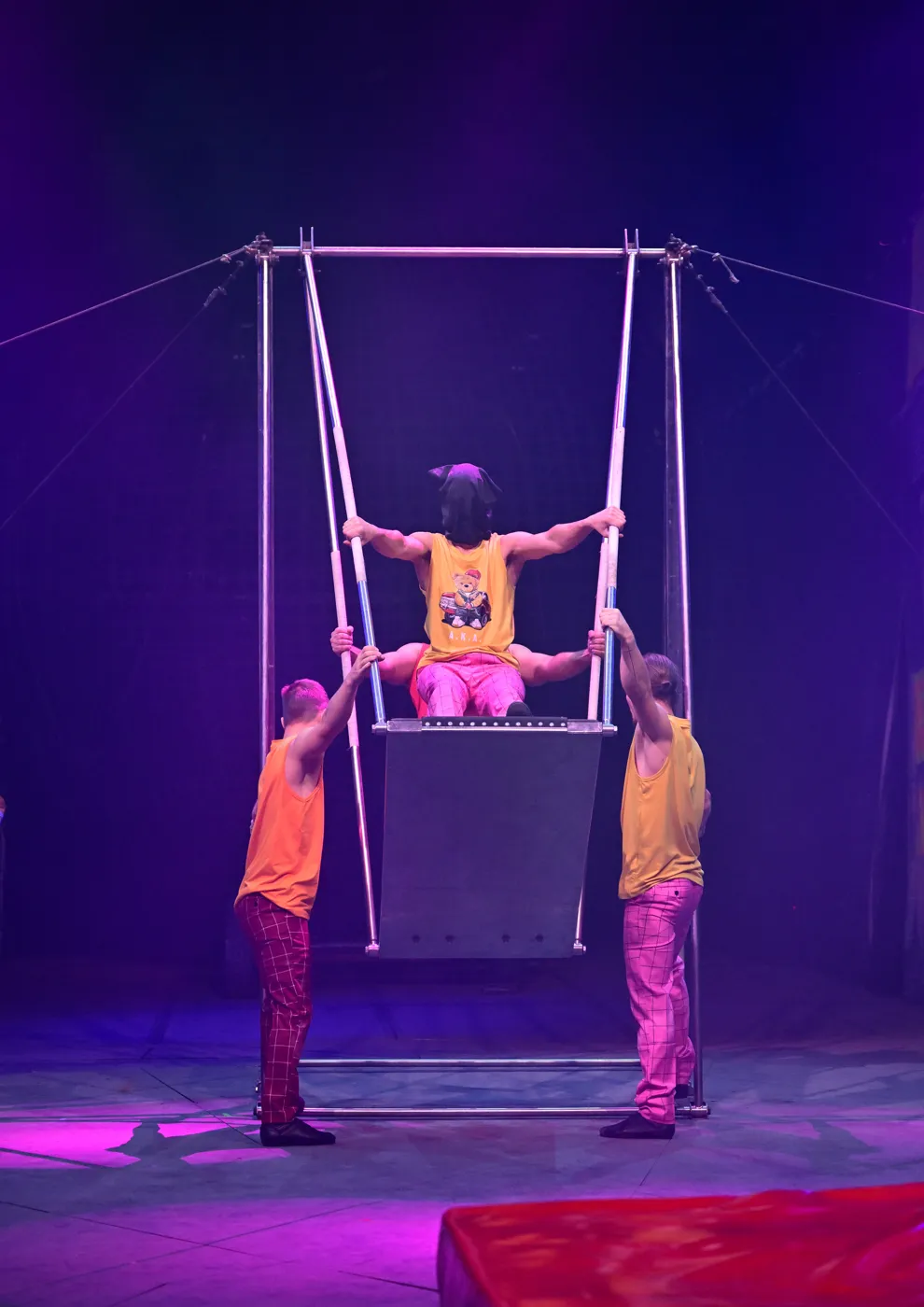 Two performers in orange steady a tall swing frame while a third sits on the platform