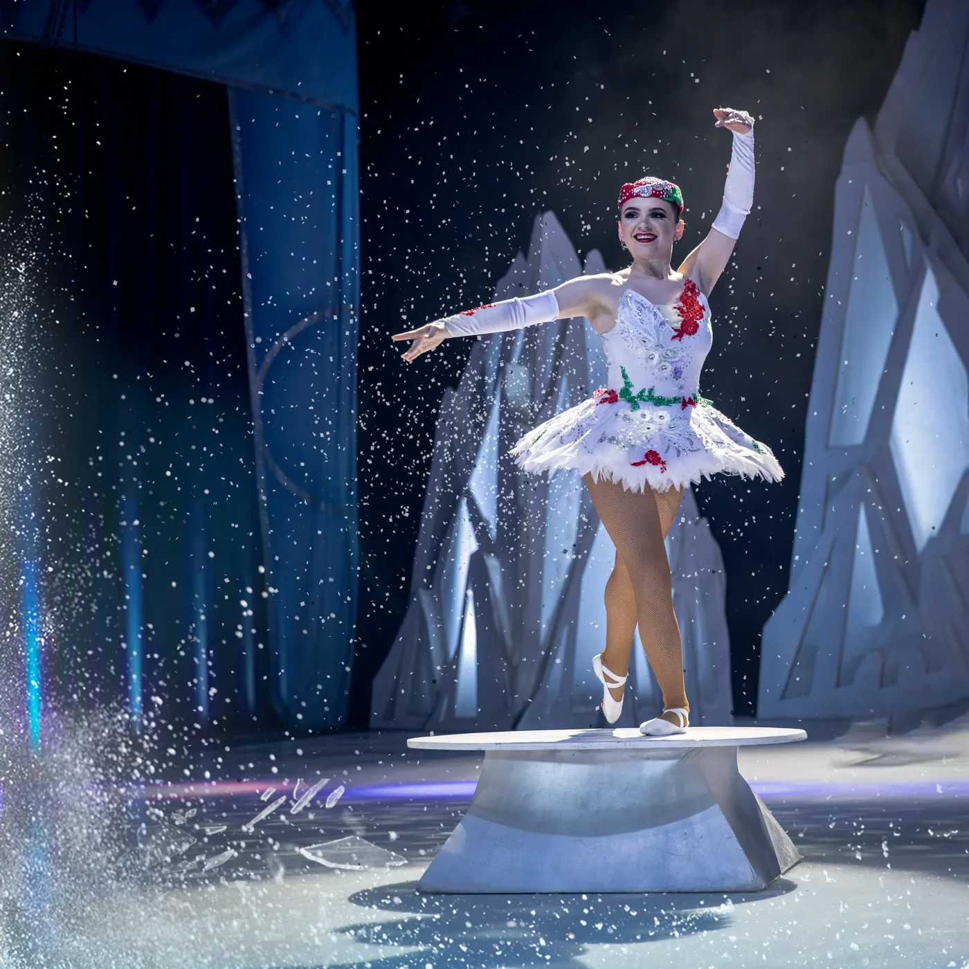 A skater in a white tutu poses on a pedestal amid falling snow