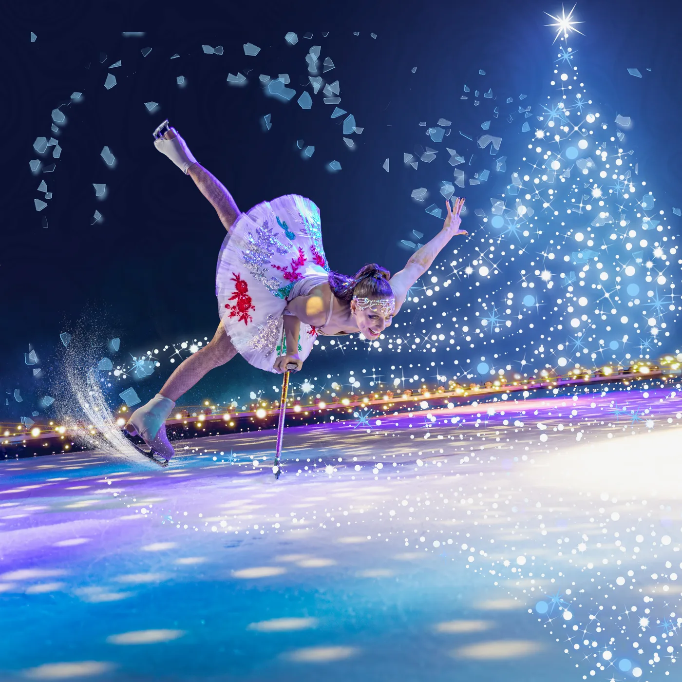 A skater in a white dress leans low on one blade beside a tree of lights