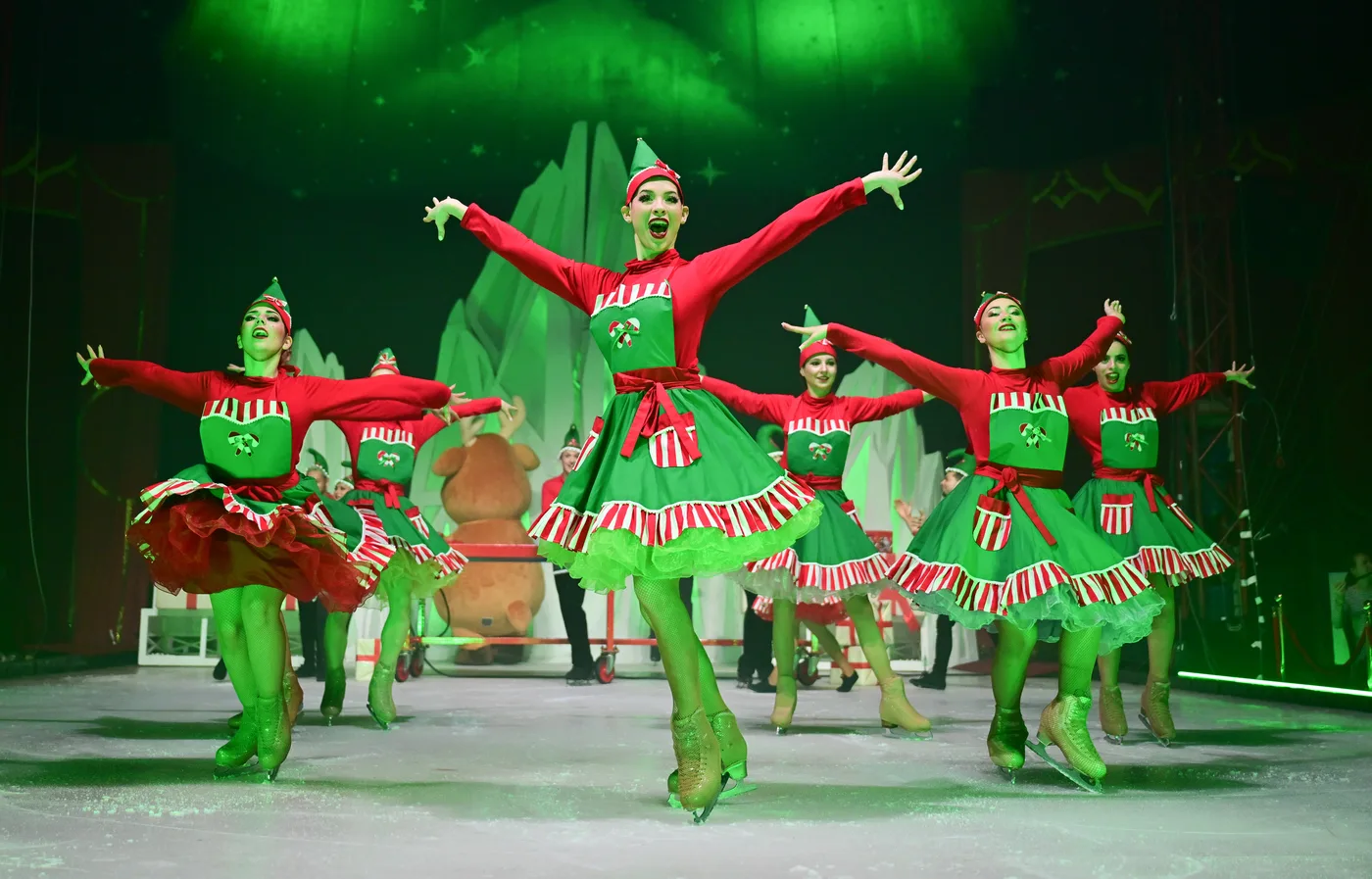 Skaters in green and red elf costumes under green lights, arms outstretched